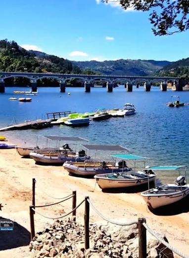 Praia fluvial na albufeira da Caniçada, com barcos e a ponte ao fundo